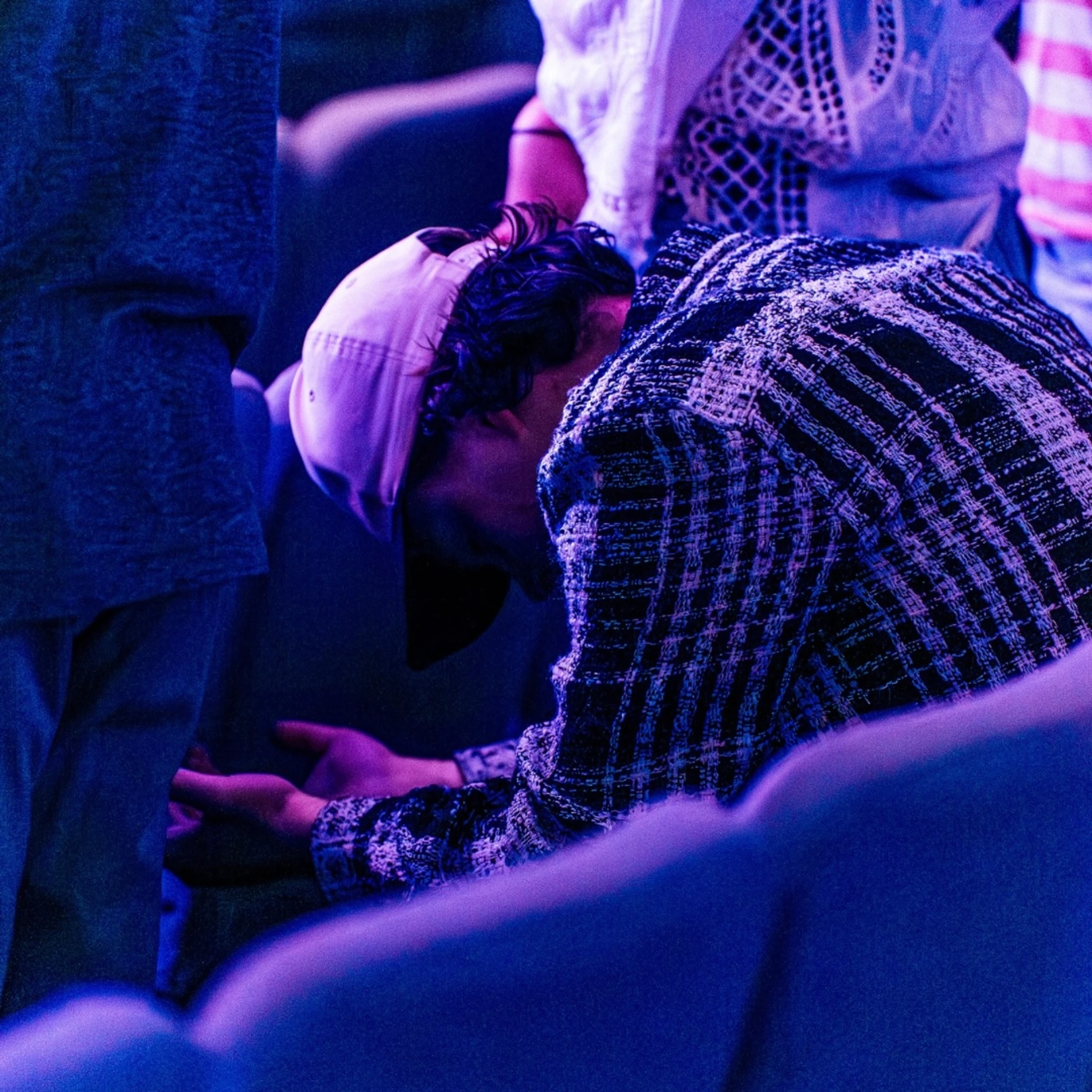 A young man in a plaid jacket and backward cap kneels between pews with his head bowed and hands open in prayer under purple and blue light.