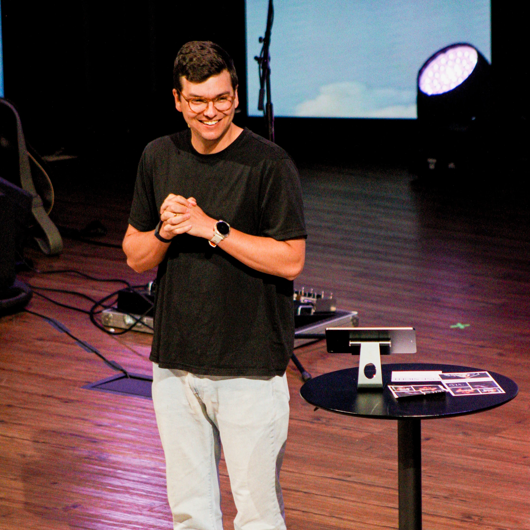 A man in a black t-shirt and glasses smiles while standing on a wooden stage. His hands are clasped, and a small table with a tablet is next to him.