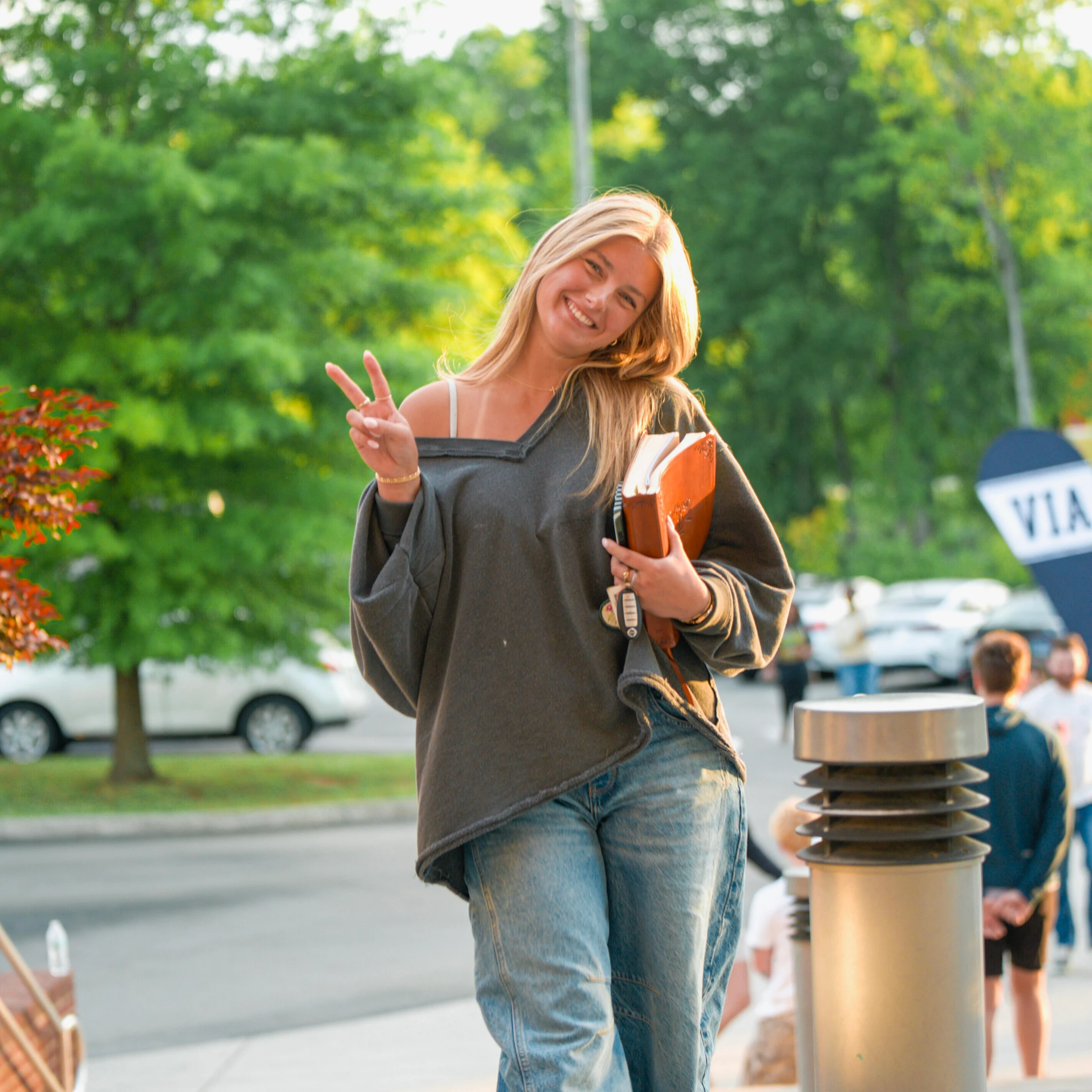 A young blonde woman in a grey sweatshirt and jeans smiles while making a peace sign. She holds a book and stands outside during the golden hour.