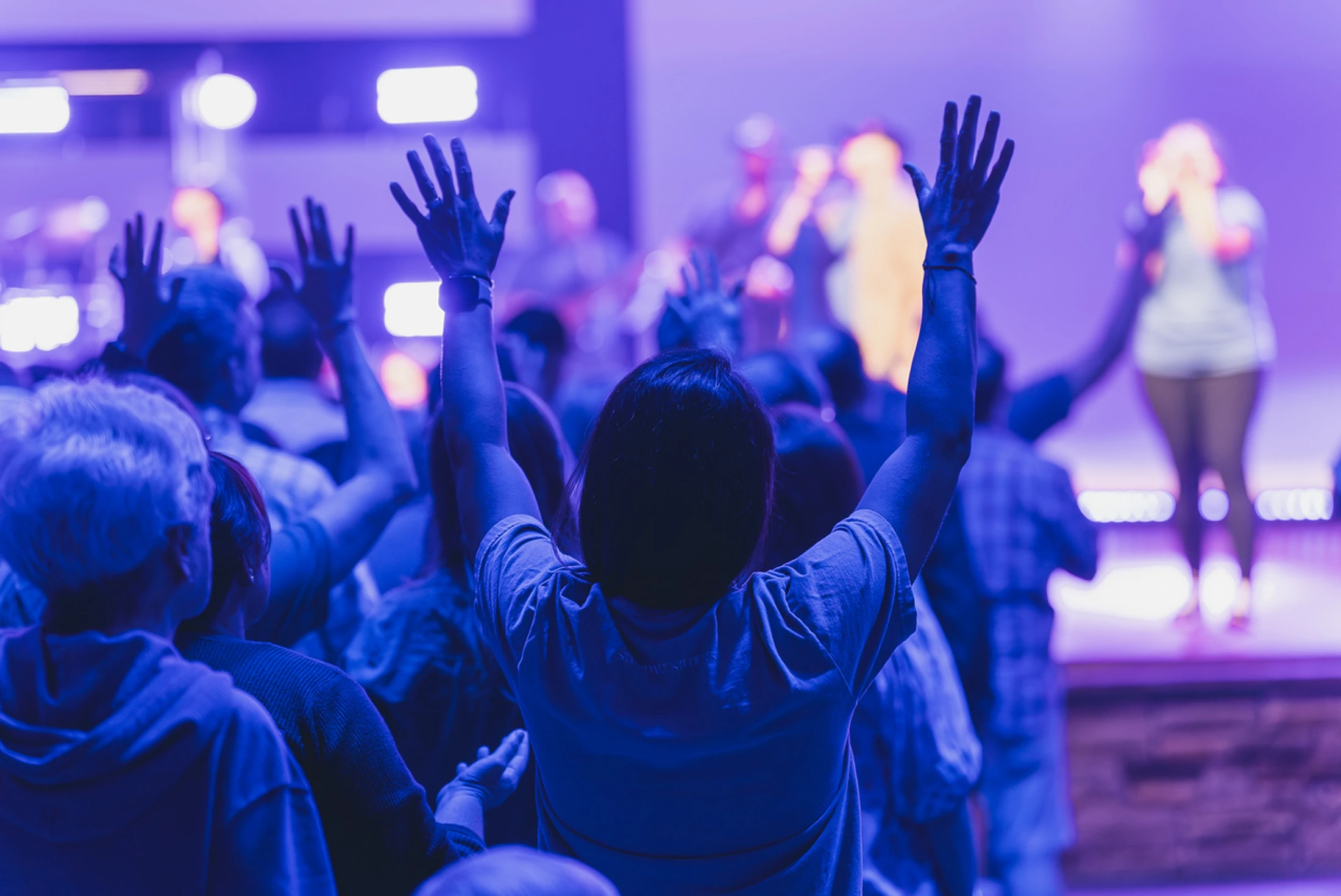 View from behind a congregation with their hands raised in worship during a church service. The scene is bathed in vibrant purple light from the stage.