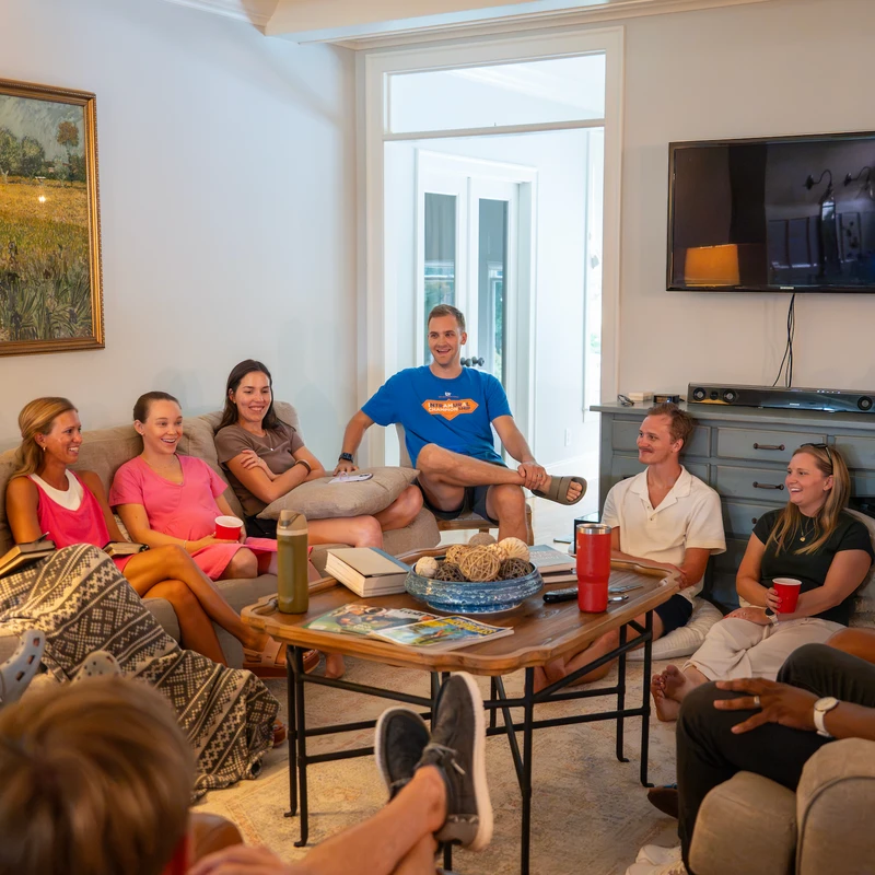 A diverse group of friends sits comfortably on couches in a bright living room, engaged in conversation and smiling during a casual social gathering.