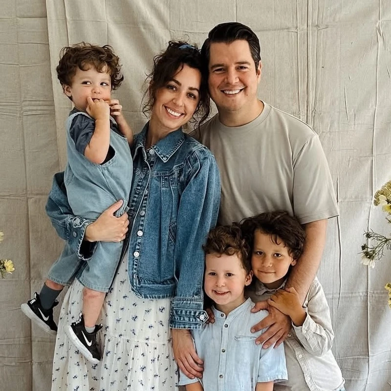 A family of five—a mother, father, and three young boys—smile for a portrait against a neutral backdrop. The mom holds the youngest child.
