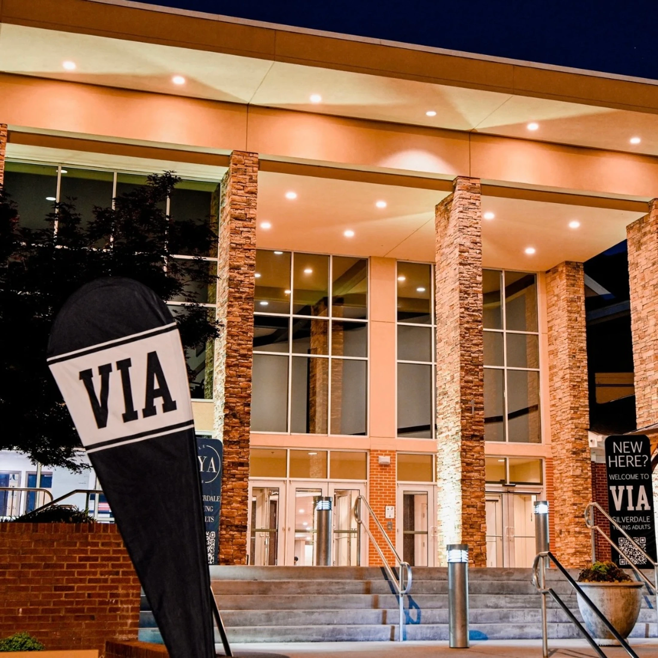 The well-lit entrance to a modern building at night, featuring large windows, stone pillars, and welcome banners for a young adults group named VIA.