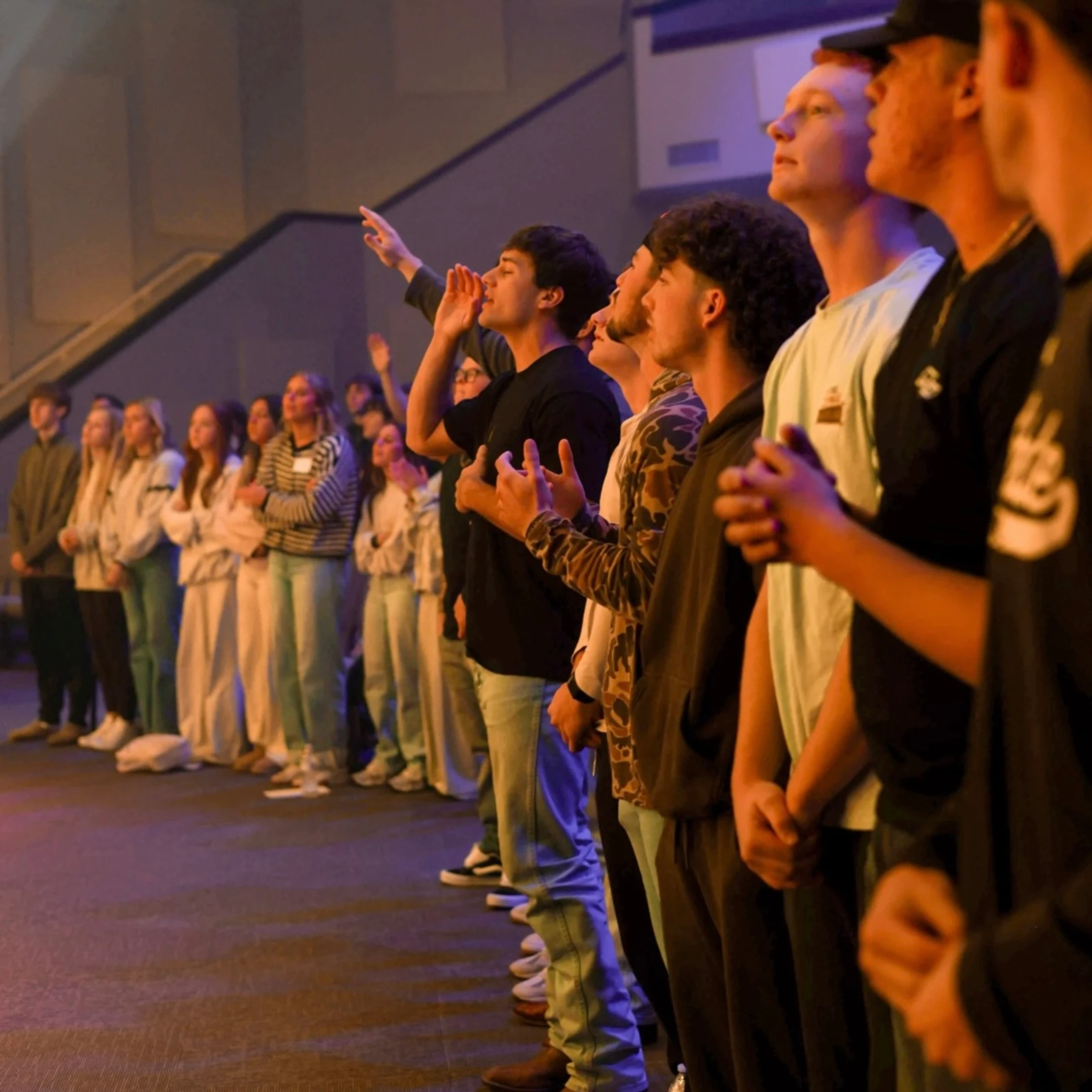 A long line of young adults stand side-by-side in a softly lit room, looking up with expressions of worship, some with hands raised.