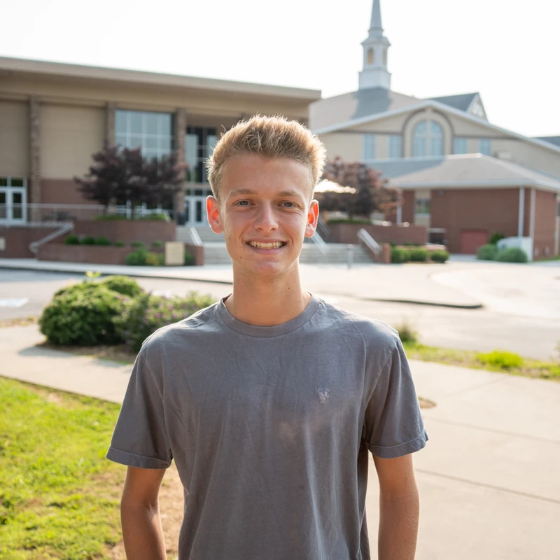 A young man with short blonde hair and a gray t-shirt smiles for the camera. He stands on a sidewalk with large buildings and trees in the background.