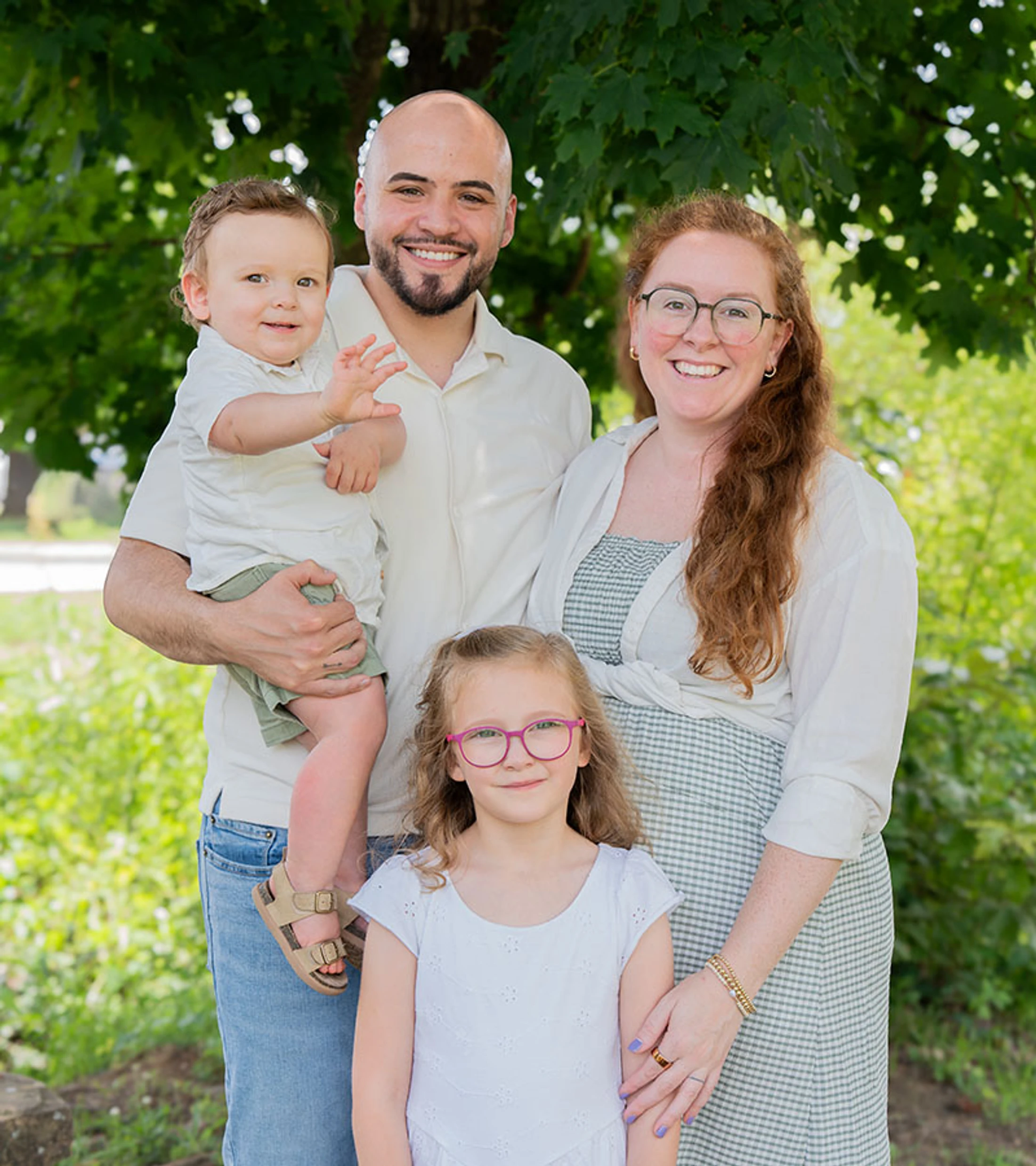 A family of four, including a mother, father, young daughter, and toddler son, smile for a portrait outdoors in front of a large green tree.