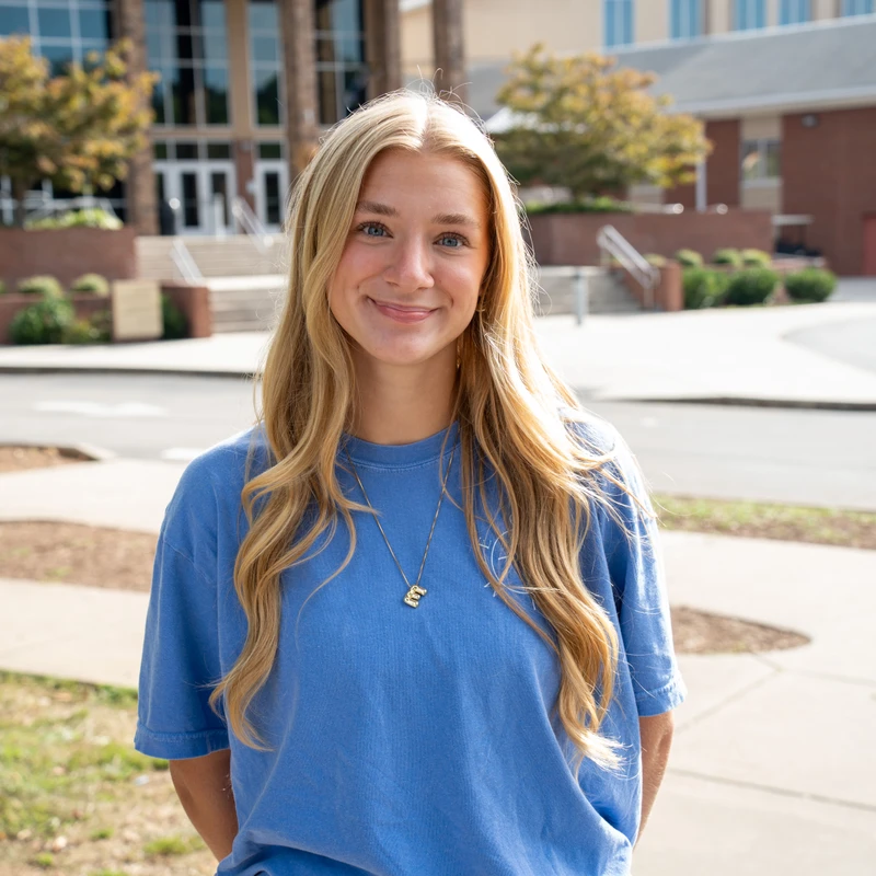 A young woman with long blonde hair and blue eyes smiles gently at the camera while standing outdoors in front of a large brick building on a sunny day.