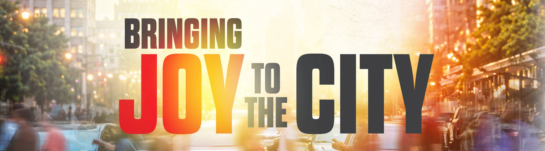 A graphic banner with the words 'Bringing Joy to the City' over a blurry, warm-toned image of a bustling city street with lights and traffic.