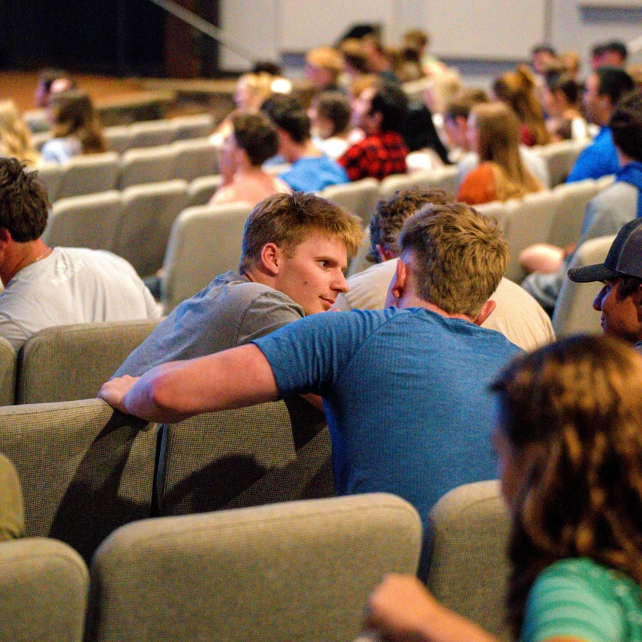 A young man in a grey t-shirt leans over his seat to talk to another young man in a blue shirt, seated in a crowded auditorium during an event.