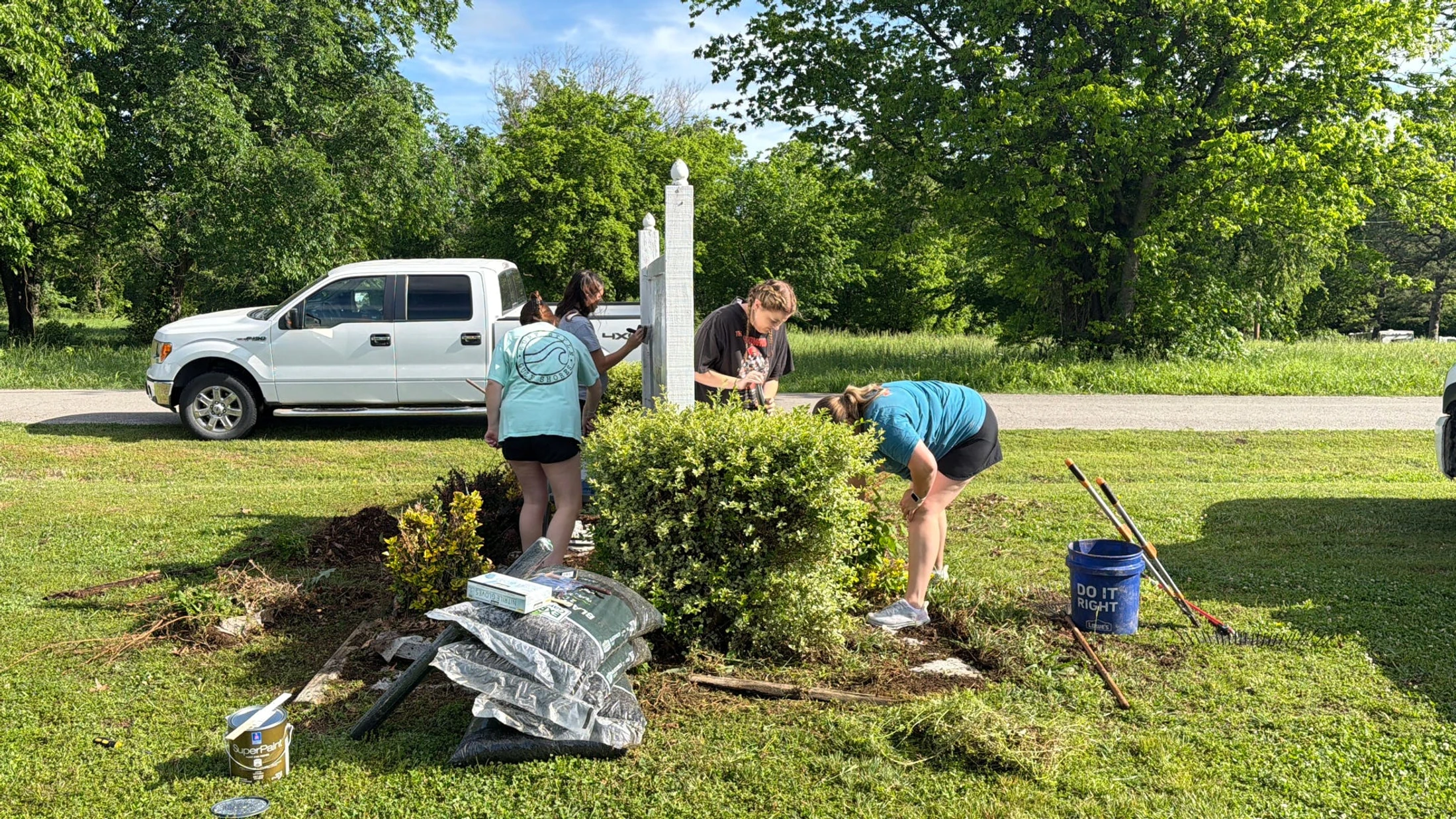 A group of four young people work on a landscaping project on a sunny day. They are painting a white post and tending to bushes, with tools nearby.