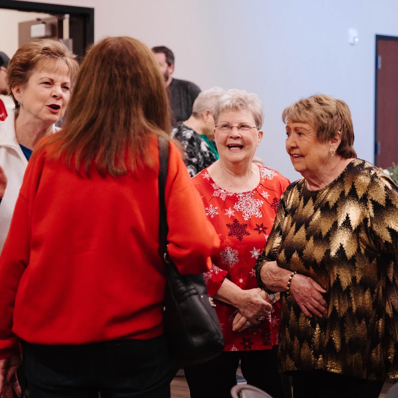 A group of older women wearing festive attire stand together talking and laughing at an indoor party with tables set up in the background.