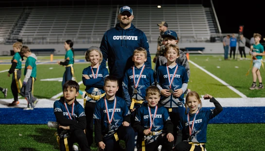 A youth flag football team called the Cowboys and their coach pose for a group photo on a turf field at night. The smiling children are all wearing medals.