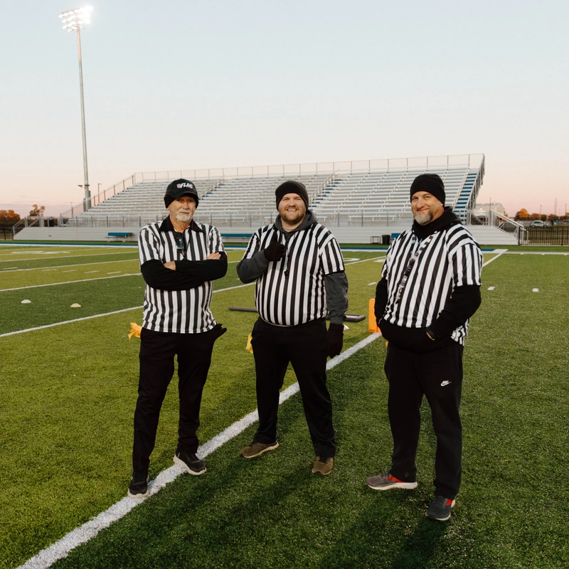 Three male football referees in black and white striped uniforms stand together on an empty football field, smiling and posing for the camera at dusk.