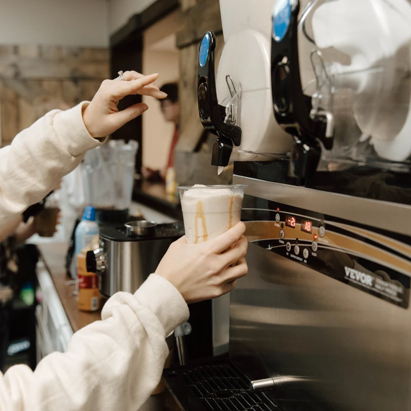 A person in a cream sweatshirt dispenses a thick, light-colored drink into a clear plastic cup from a stainless steel beverage machine in a cafe.