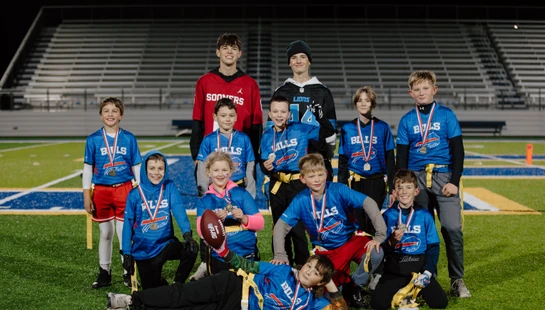 A youth flag football team in blue jerseys poses for a group photo on a turf field at night. The children are smiling and many are wearing medals.