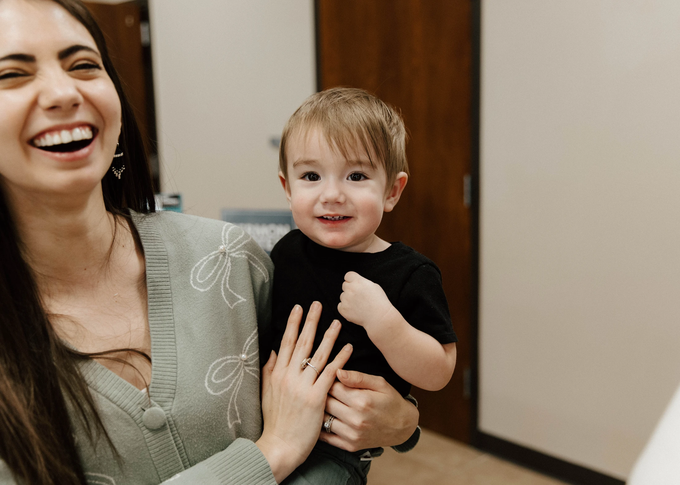 A happy woman with long dark hair laughs while holding a small boy in a black t-shirt who is smiling at the camera.