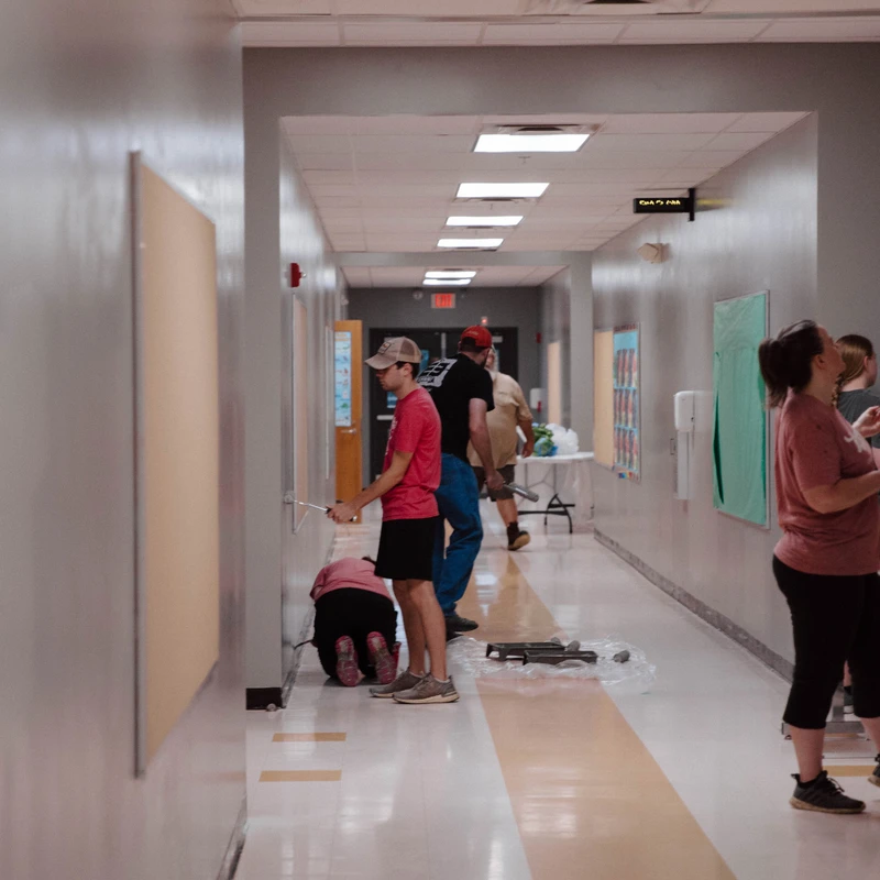 A group of volunteers of various ages paint the grey walls of a long school corridor with rollers and brushes during a community project.