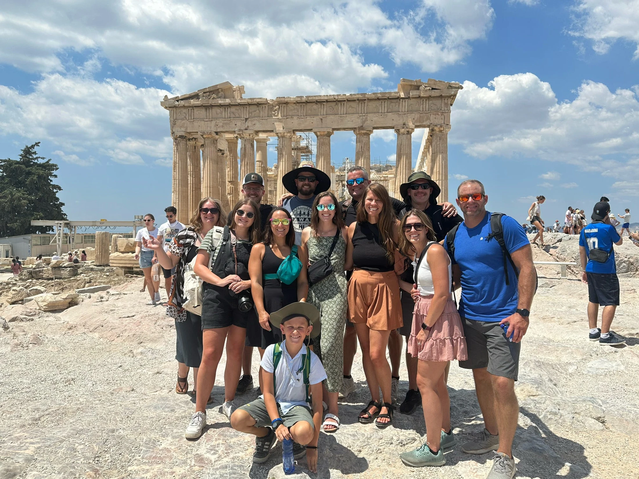 A large group of smiling tourists posing for a photo in front of the Parthenon at the Acropolis in Athens on a sunny day with blue skies.