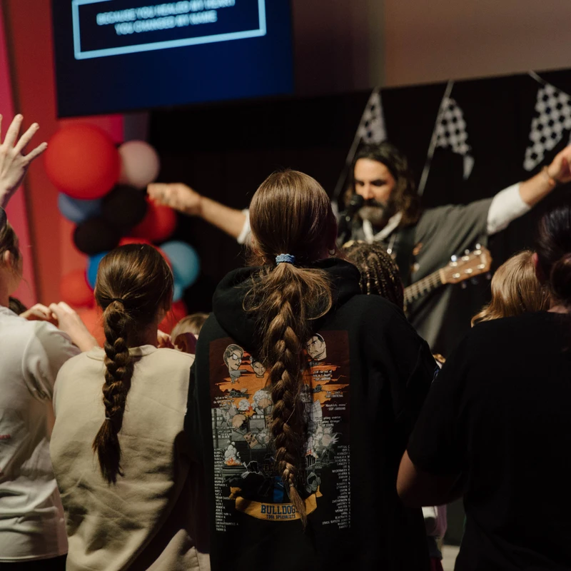 A view from behind a crowd of young people facing a man on stage with a guitar. Some have their hands raised in worship during a church service or concert.