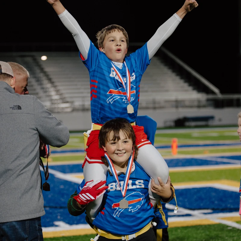 A young boy sits on his teammate's shoulders, arms raised in victory. Both wear blue football jerseys, medals, and stand on a field at night.