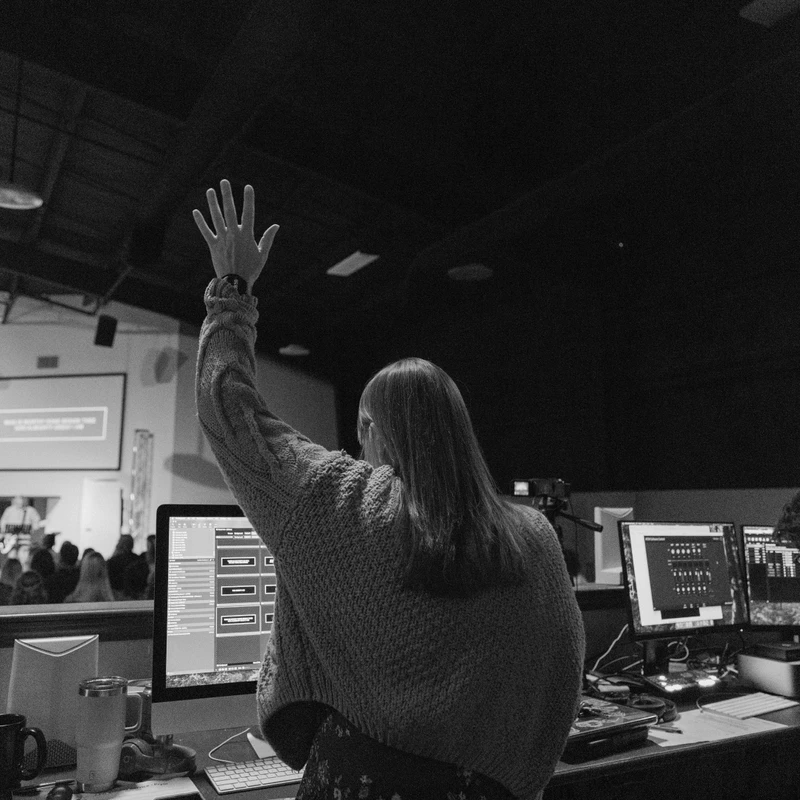 A black and white photo from behind a woman in a sweater raising her hand in worship while working at a tech booth during a church service.