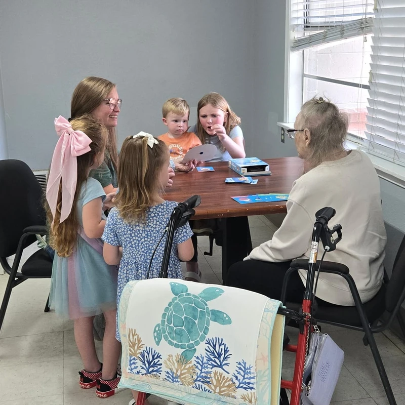 A group of young children and a woman gather around a table with an elderly woman, playing a card game. A red walker is in the foreground.