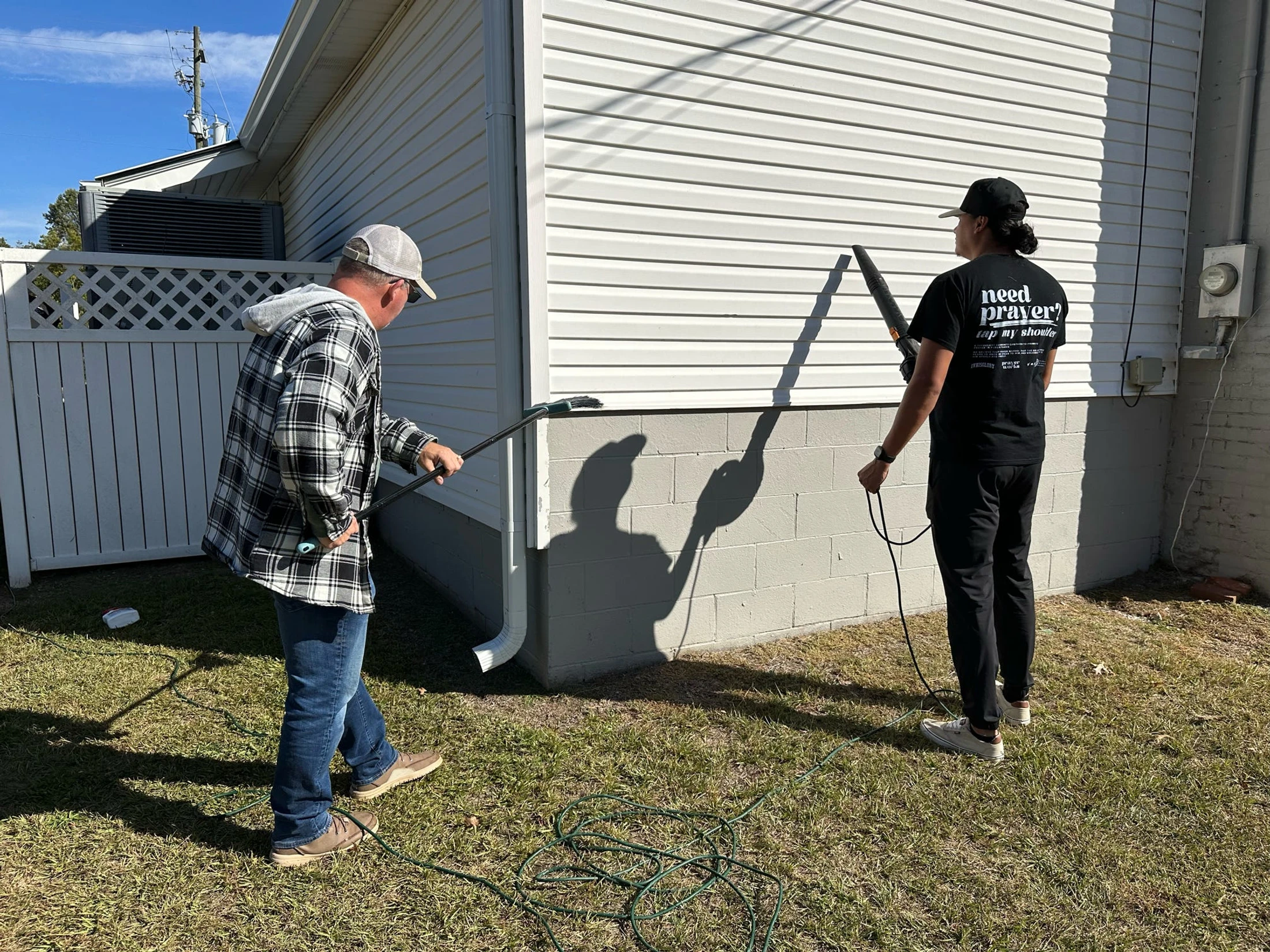 Two men work together to clean the exterior of a house. One uses a long brush on the siding while the other holds a leaf blower on a sunny day.