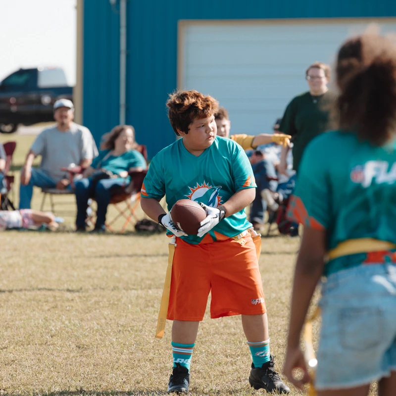 A young boy in a turquoise and orange jersey holds a football during a youth flag football game on a grassy field, with other players and spectators nearby.