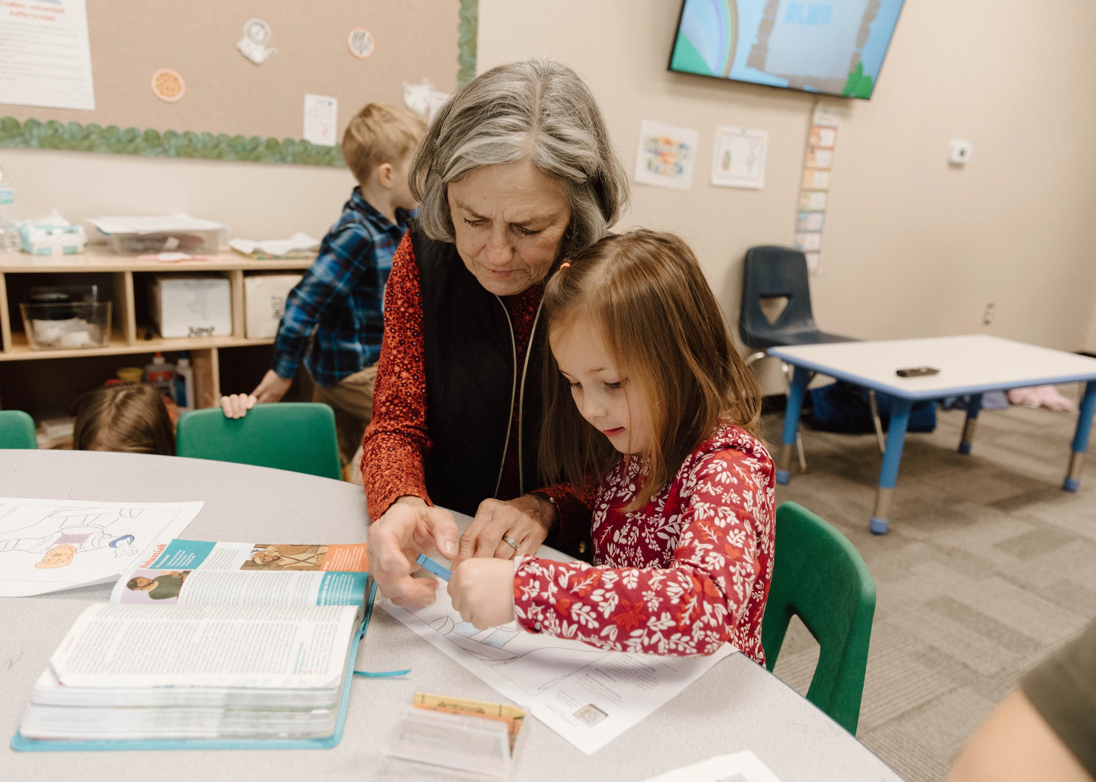 An older woman with gray hair leans over to help a young girl in a red floral shirt as she works on a coloring page at a table in a children's classroom.