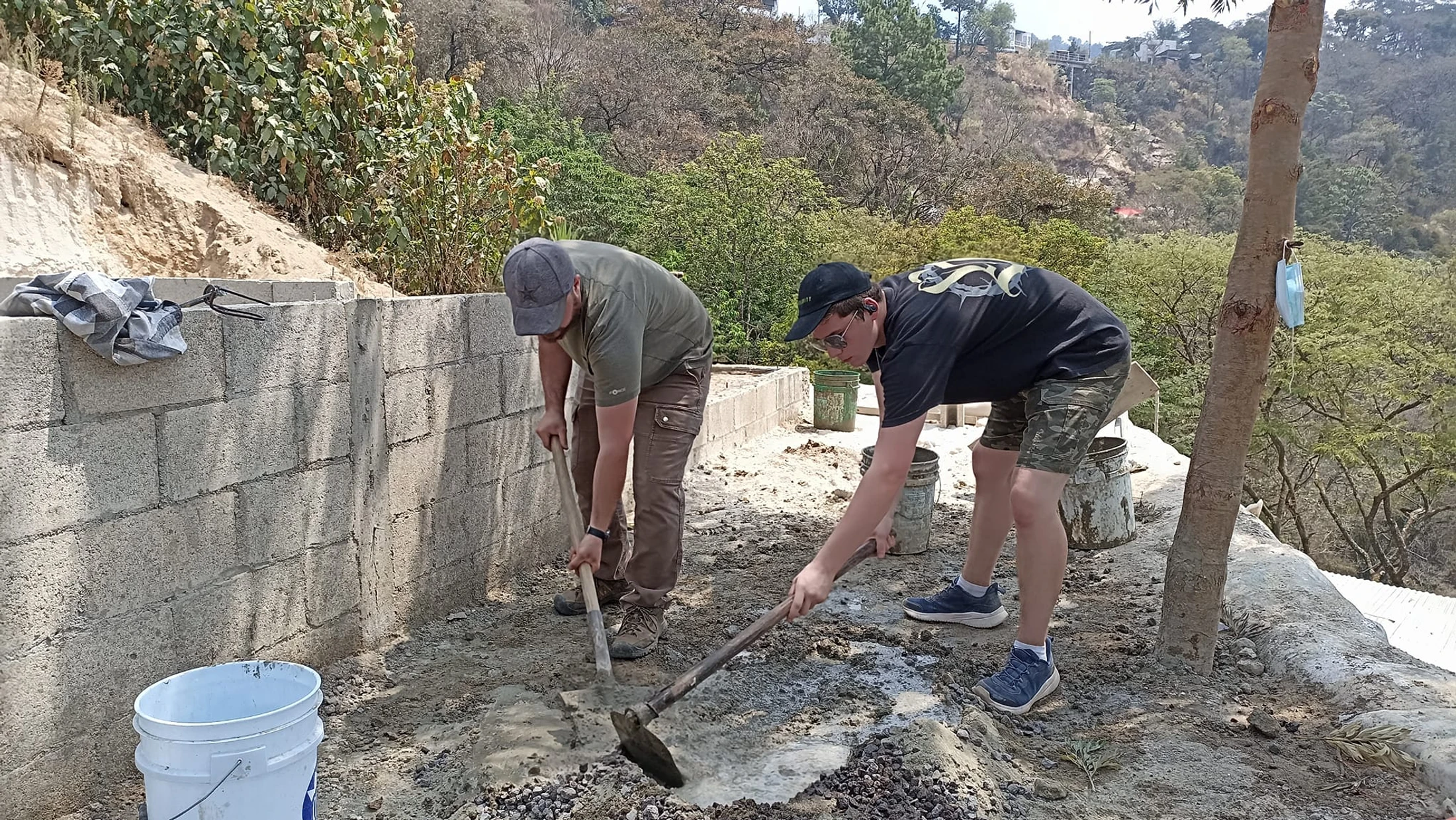 Two men in caps use hoes to mix wet concrete and gravel on the ground next to a cinder block wall, with a lush, green hillside in the background.