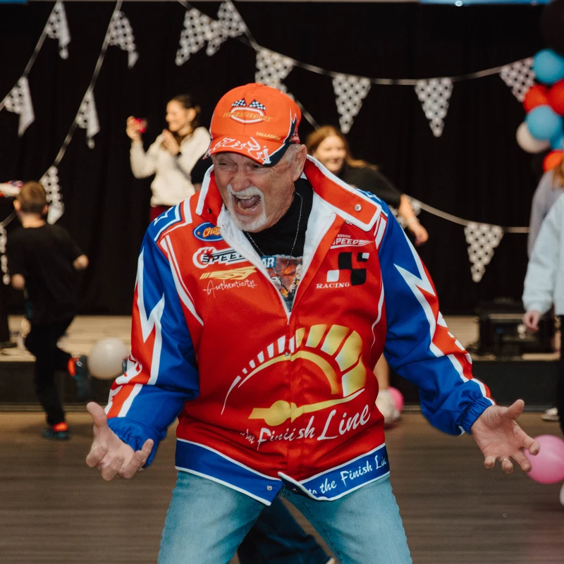 An older man with a white goatee and an excited expression wears a colorful racing jacket and orange cap at a party with checkered flags and balloons.