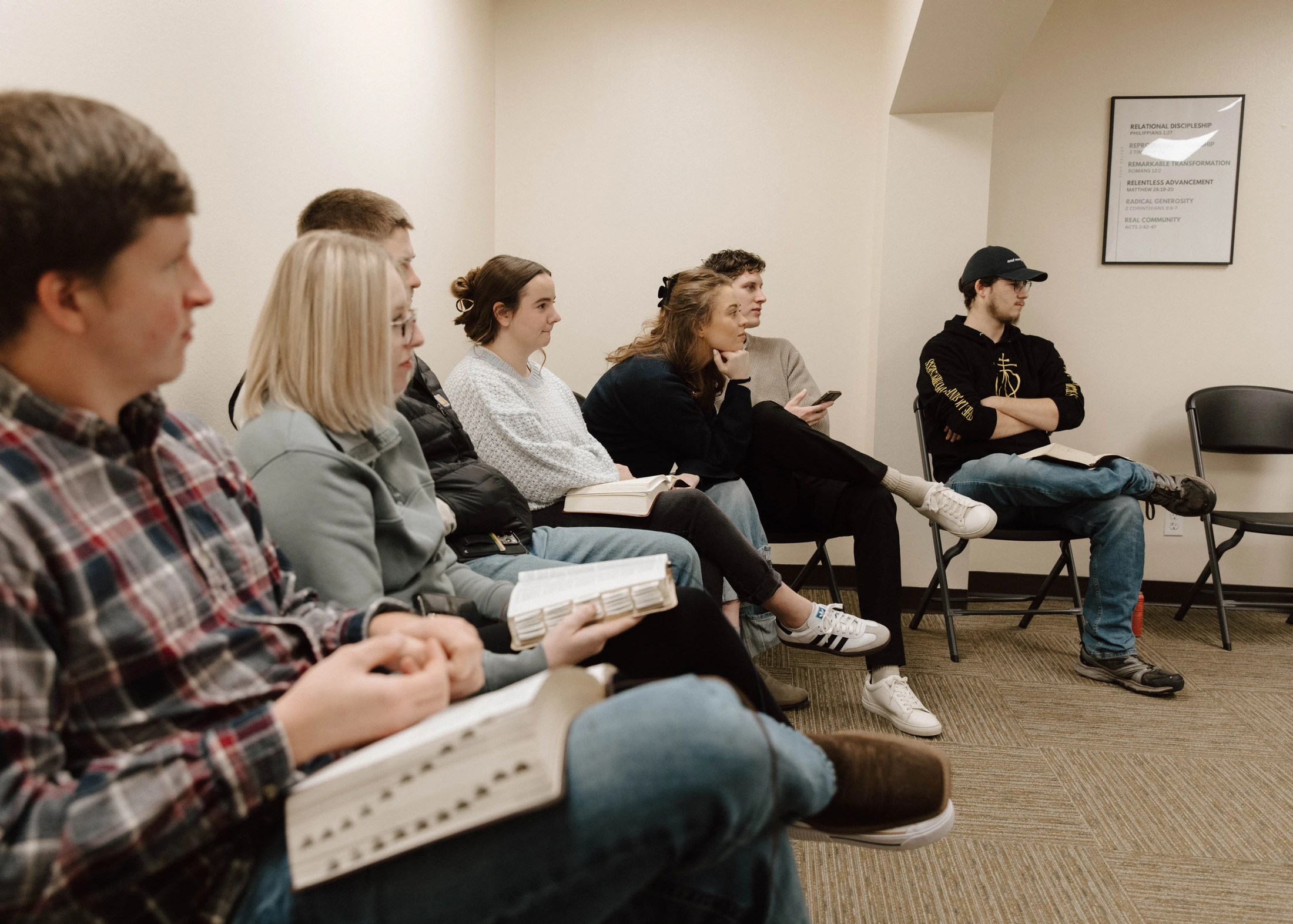 A group of young adults sits in a row of chairs, listening attentively in a meeting room. Many hold open books, likely Bibles, on their laps.