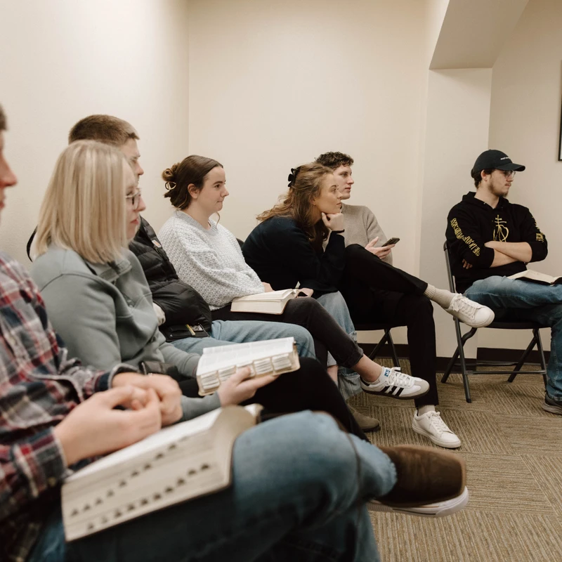 A group of young adults sits in a row of chairs, listening attentively in a meeting room. Many hold open books, likely Bibles, on their laps.