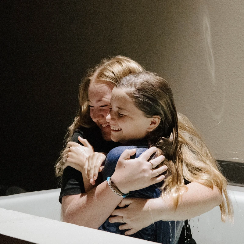 Two young women with wet hair and clothes smile and hug each other tightly while standing in a white baptismal font during a religious ceremony.