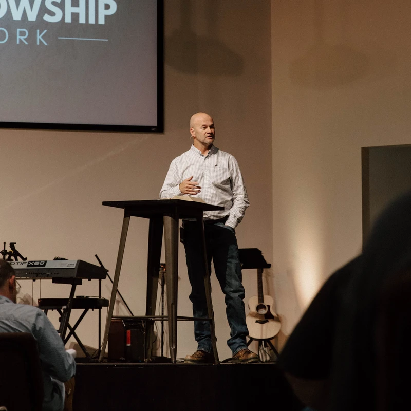 A bald man in a checkered shirt and jeans stands on a stage behind a lectern, speaking to an audience. A screen behind him says 'Fellowship Network'.
