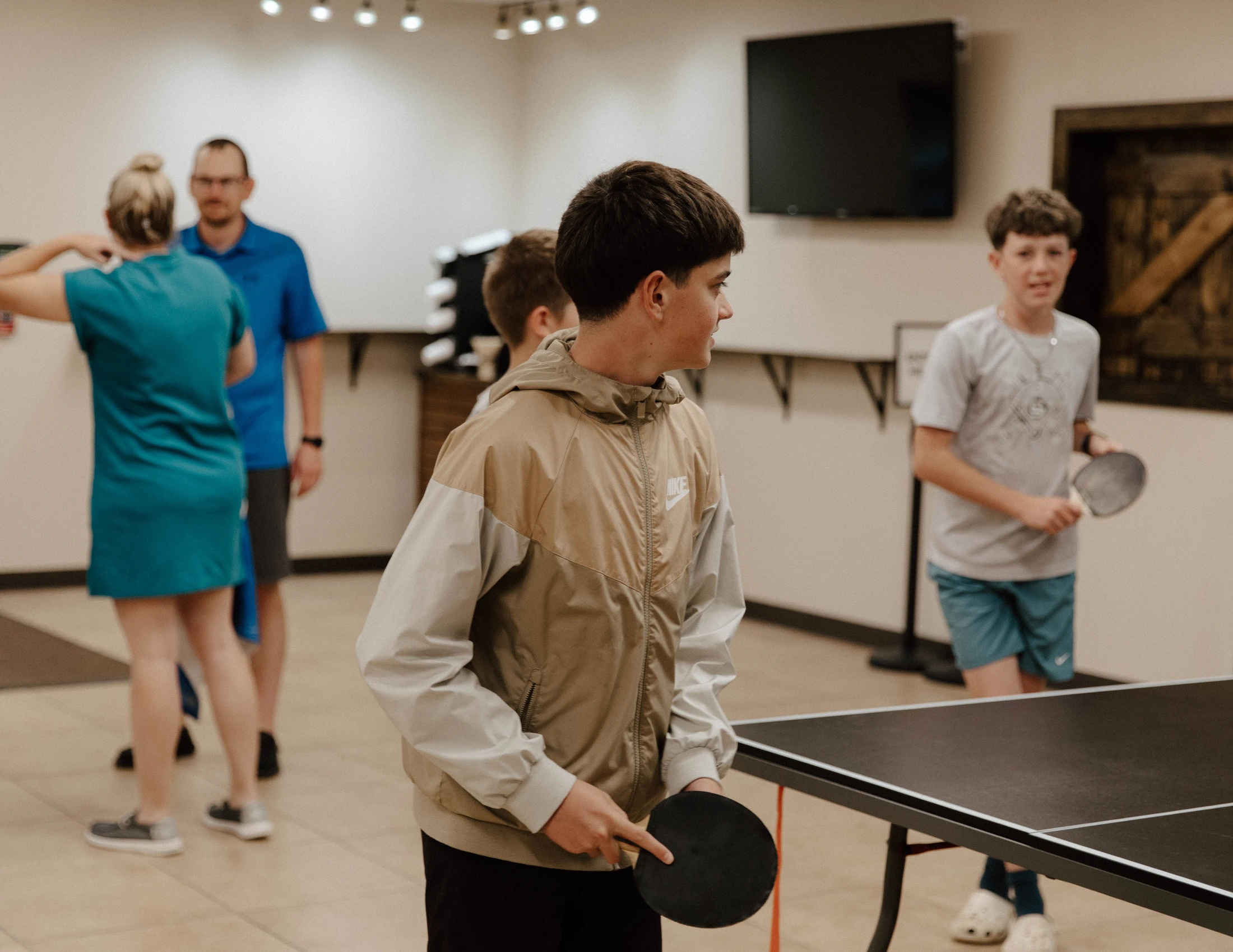Two teenage boys stand by a ping pong table holding paddles. One boy in a tan jacket is in the foreground, while adults stand talking in the background.