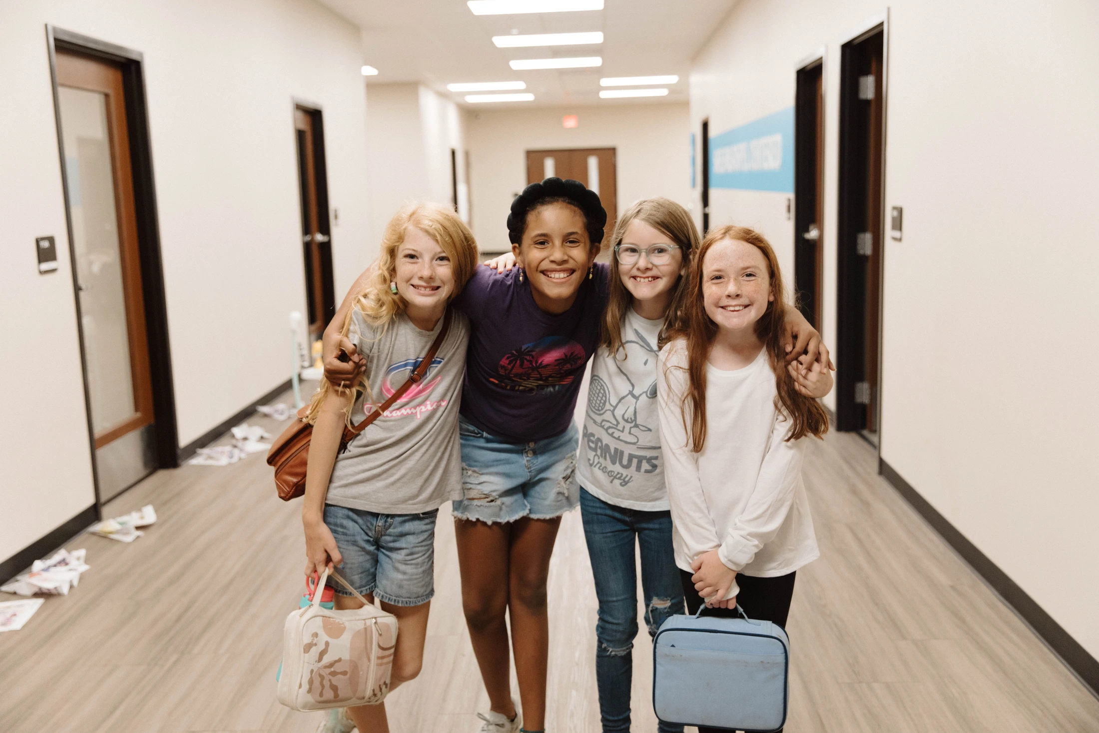 A diverse group of four young girls smile for the camera as they stand with their arms around each other in a school hallway.