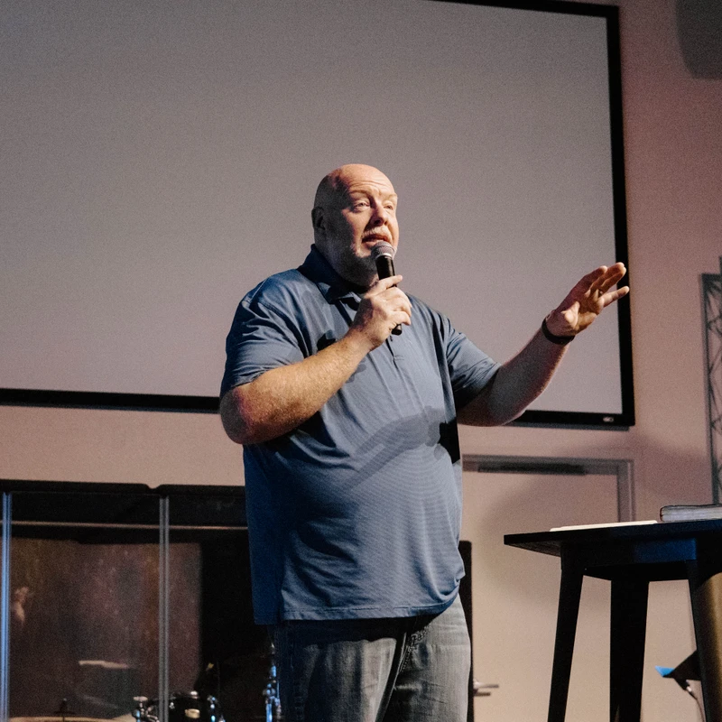 A bald man in a blue polo shirt stands on a stage, speaking into a microphone and gesturing. A large projection screen is behind him.