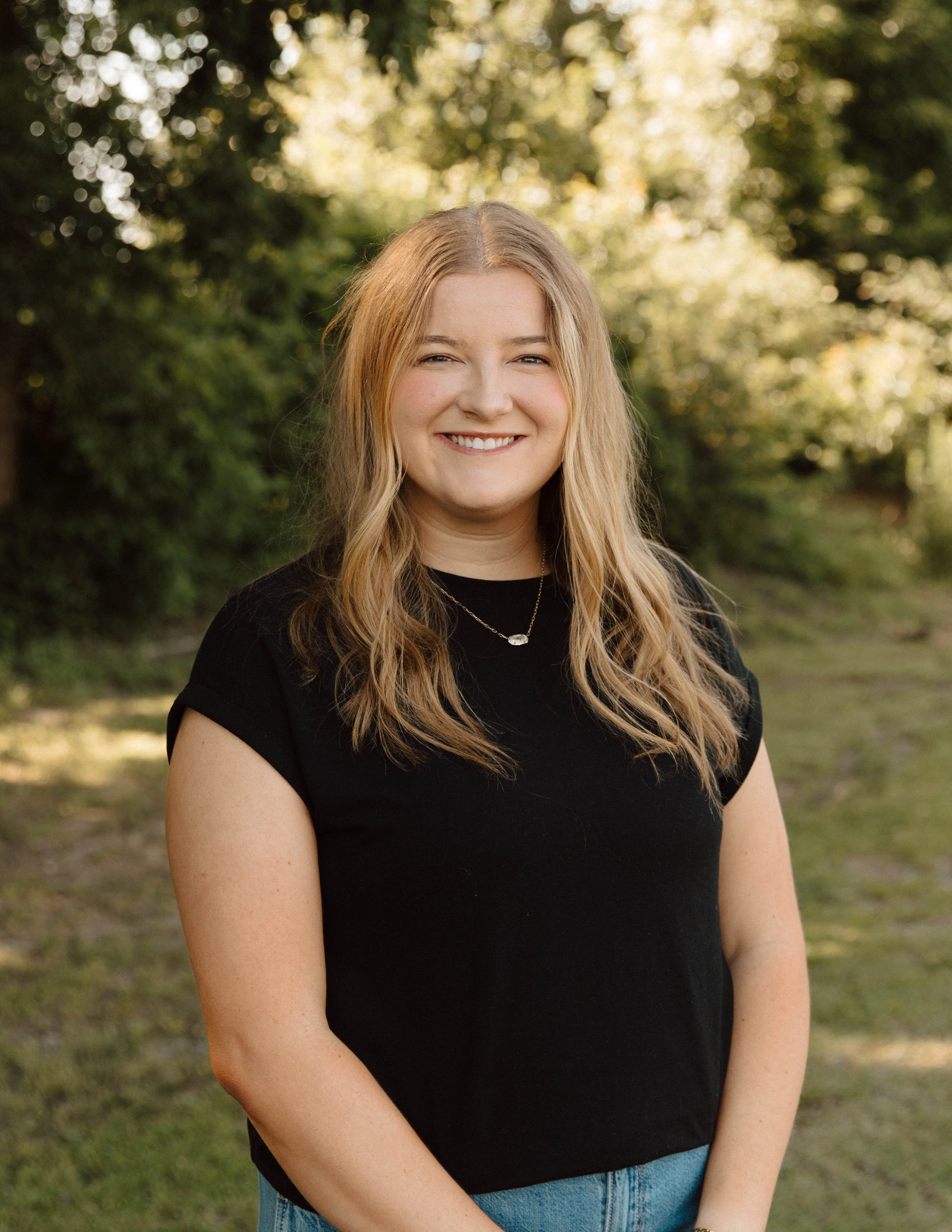 A head-and-shoulders portrait of a smiling woman with long blonde hair. She is wearing a black shirt and stands outdoors with a background of green trees.