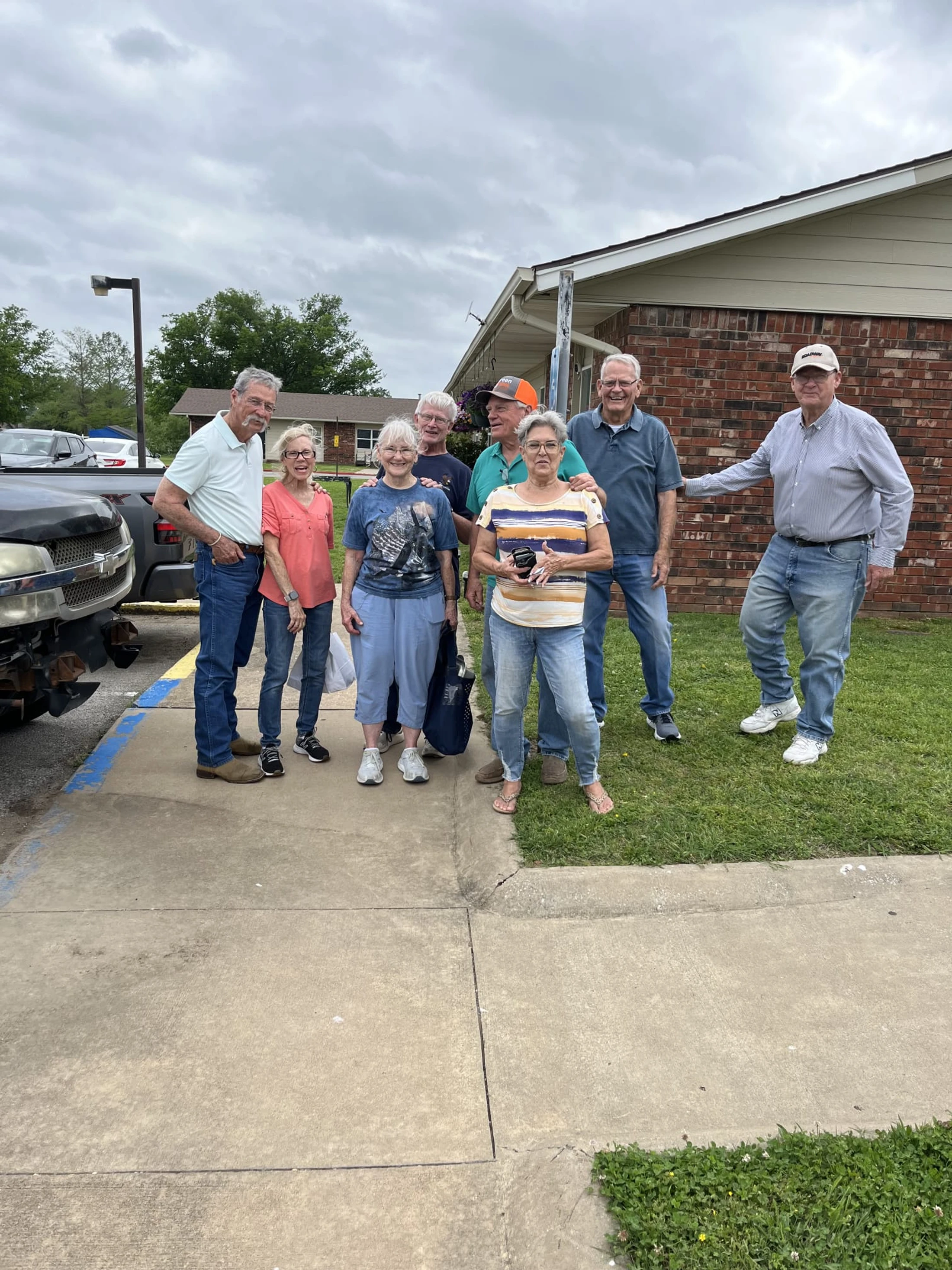 A group of eight smiling senior citizens posing for a photo on a sidewalk in front of a brick building on a cloudy day.