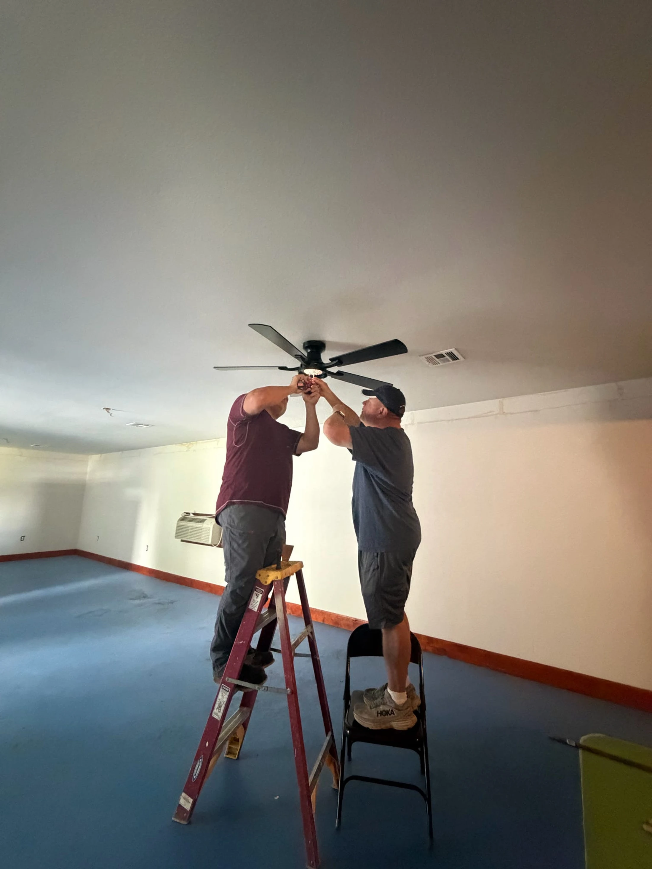 Two men, one on a ladder and another on a chair, collaborate to install a black ceiling fan in a room with a blue floor and white walls.