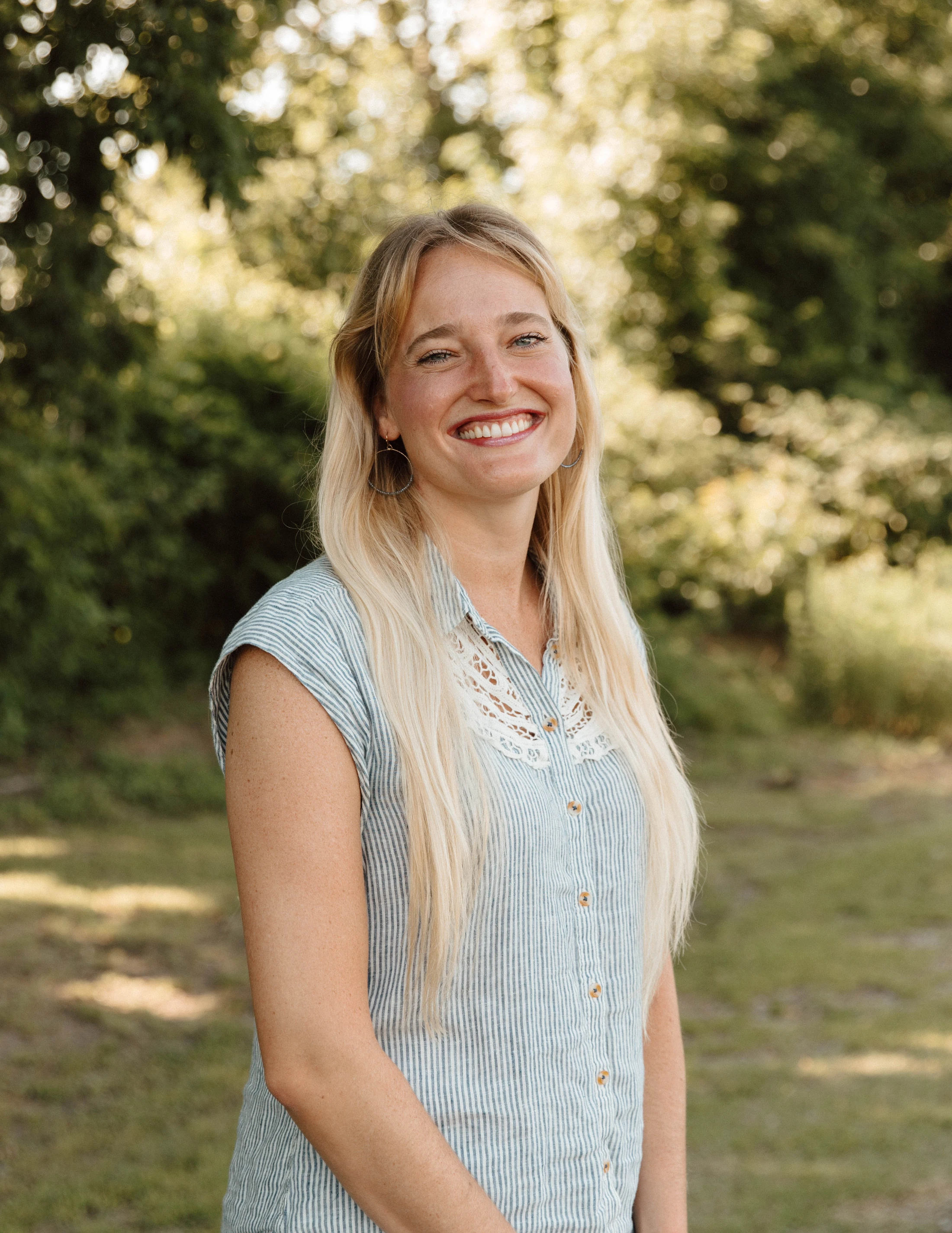 A young woman with long blonde hair and freckles smiles warmly. She is standing outdoors in a park, wearing a blue and white striped shirt with a lace collar.