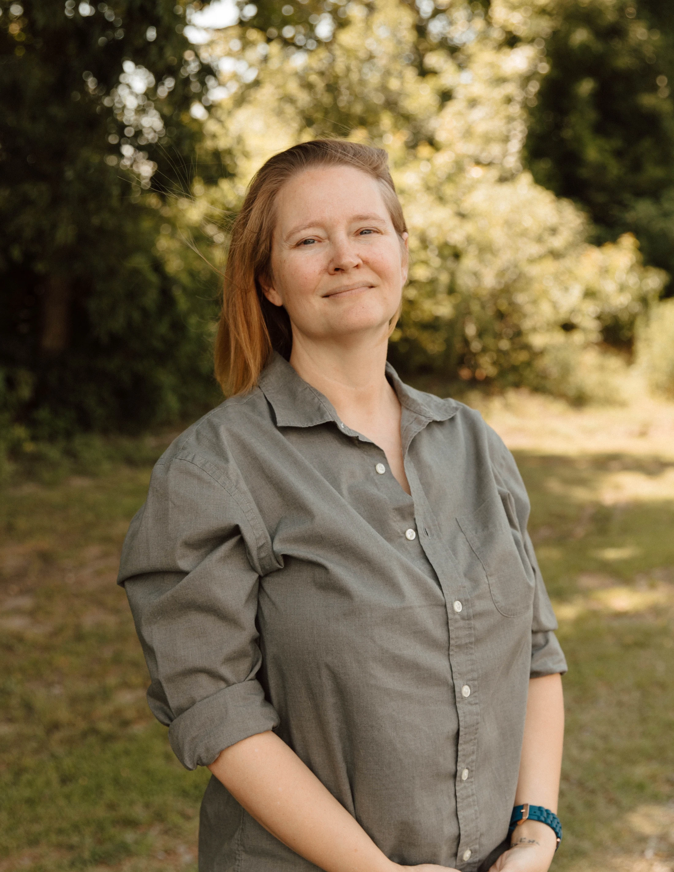 A woman with reddish-blonde hair, wearing a gray button-up shirt, smiles gently at the camera while standing outside with a blurred background of green trees.