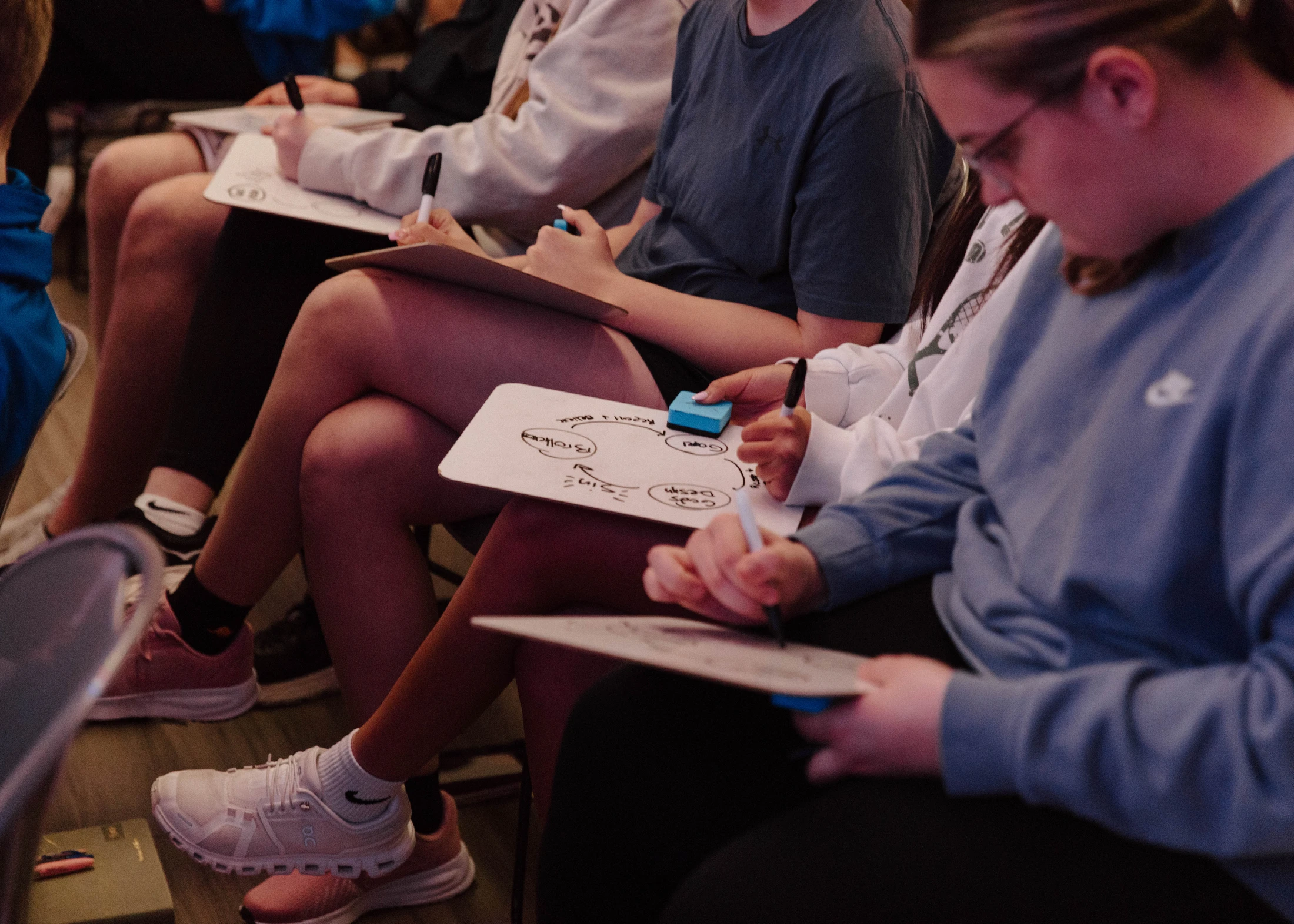 A row of seated students actively participating in a lesson, writing notes and drawing diagrams on small whiteboards resting on their laps.