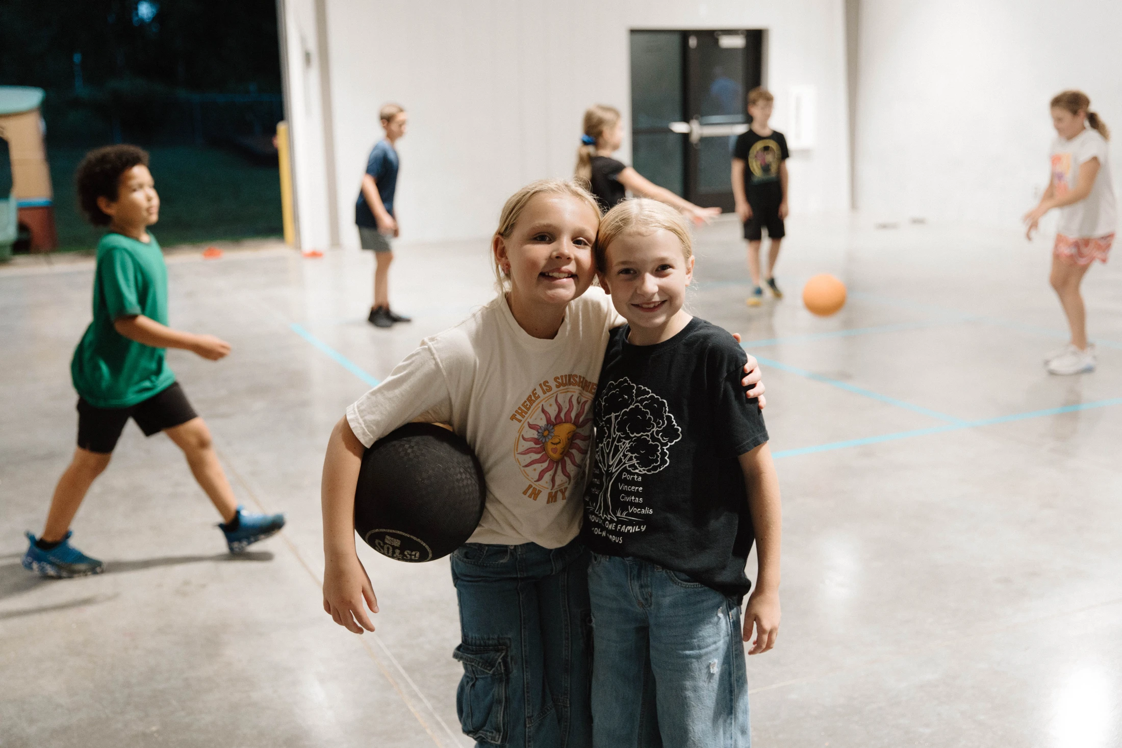 Two young girls smile and hug for a photo in a gymnasium. One holds a black ball while other children play in the background.