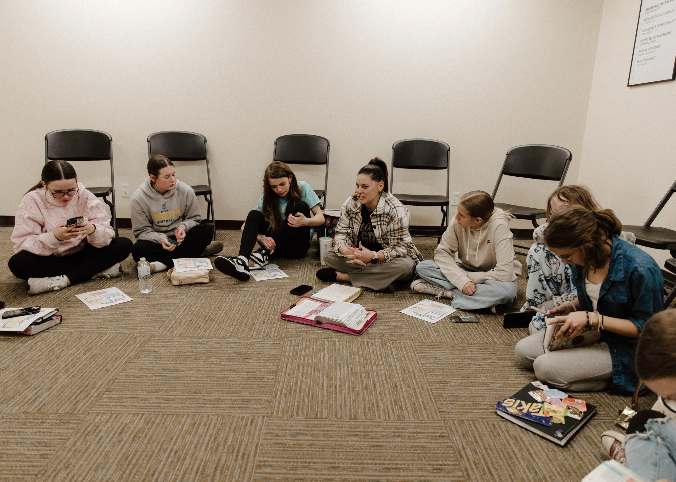 A group of young women and girls sit in a circle on a carpeted floor, participating in a bible study or small group discussion.
