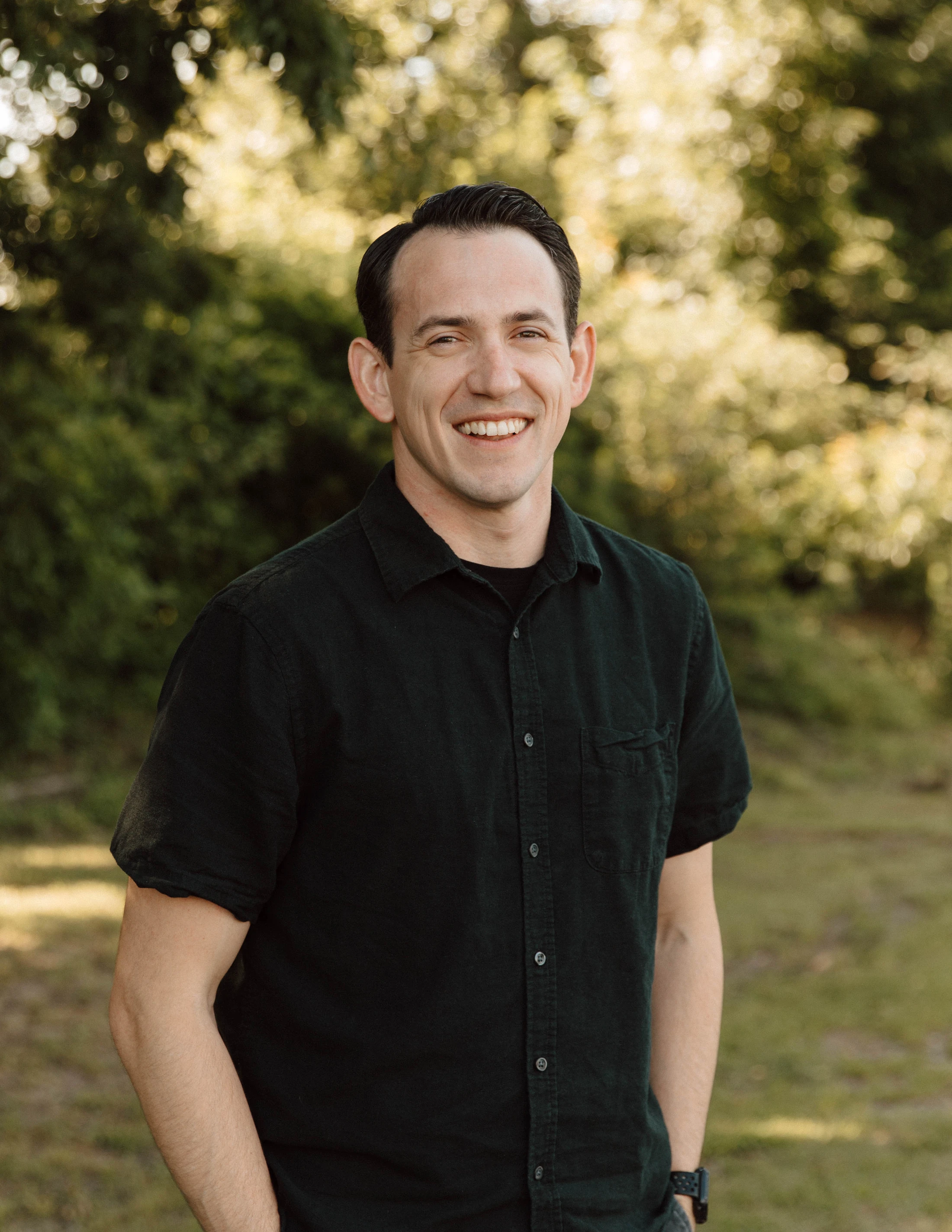 A man with short dark hair smiles warmly at the camera. He wears a dark short-sleeve button-up shirt and stands outside with a blurry green background.