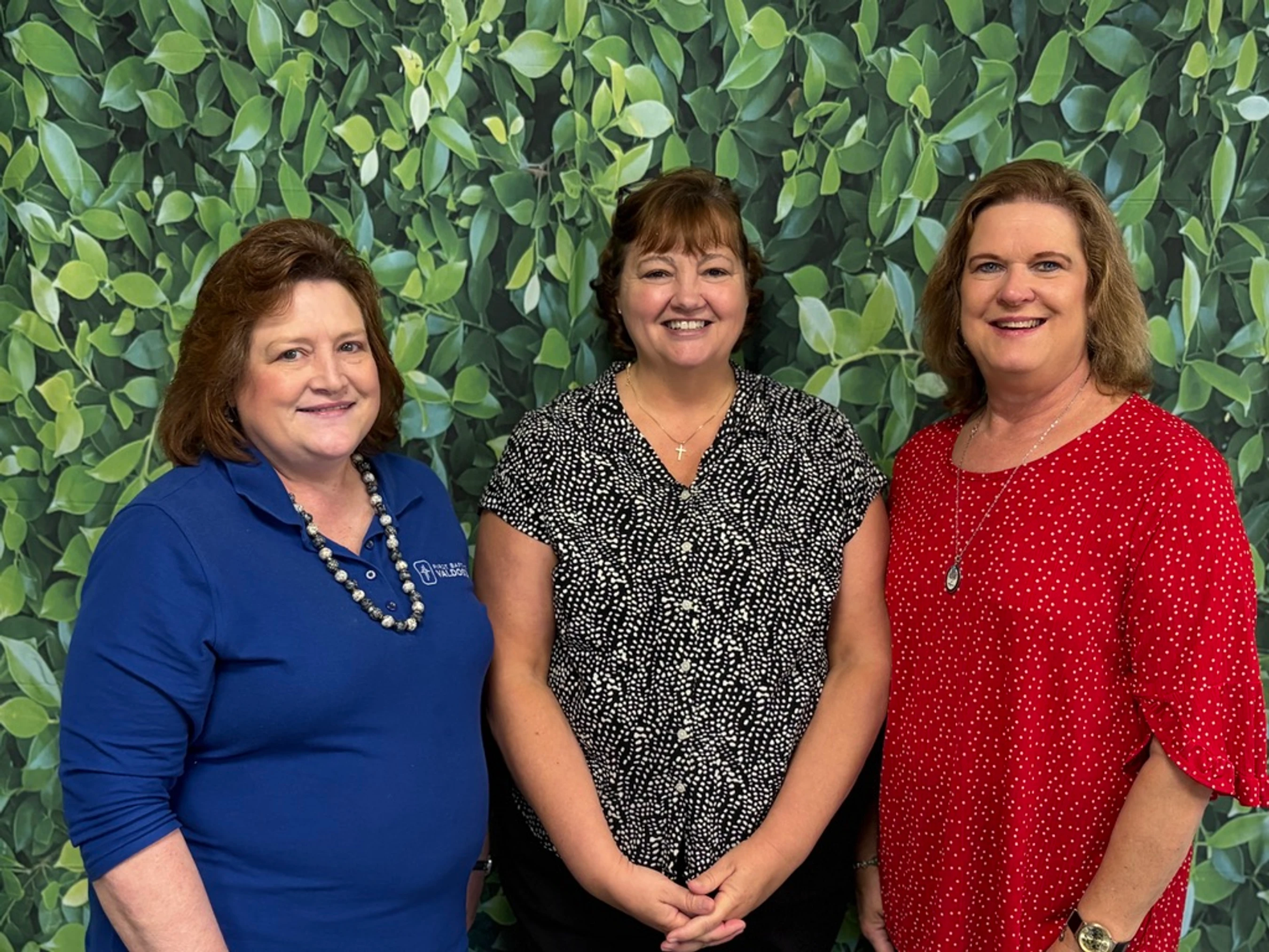 Three middle-aged women stand side-by-side smiling for a photo in front of a green leafy backdrop.