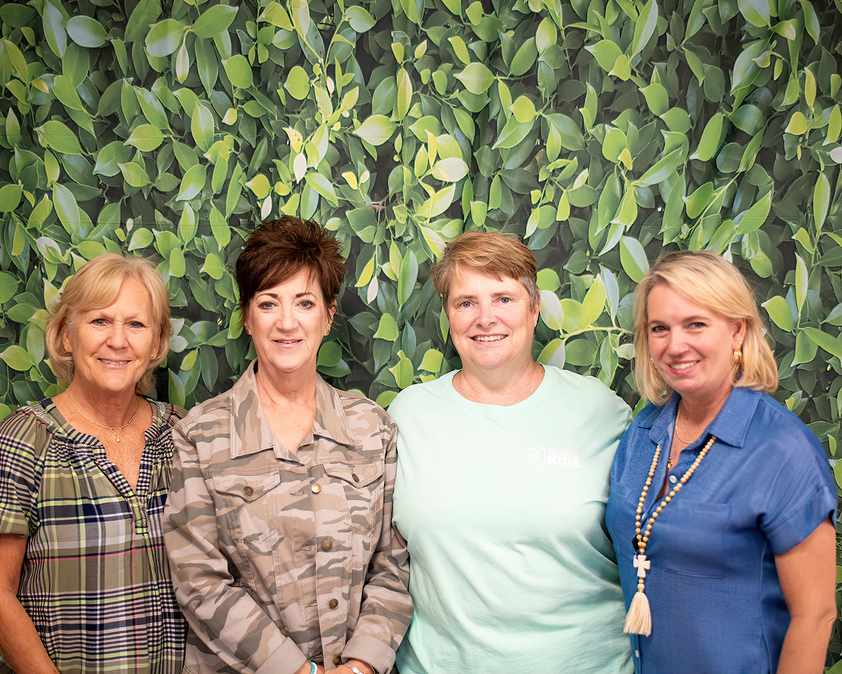 A group photo of four smiling women standing shoulder to shoulder in front of a wall with a pattern of green leaves.