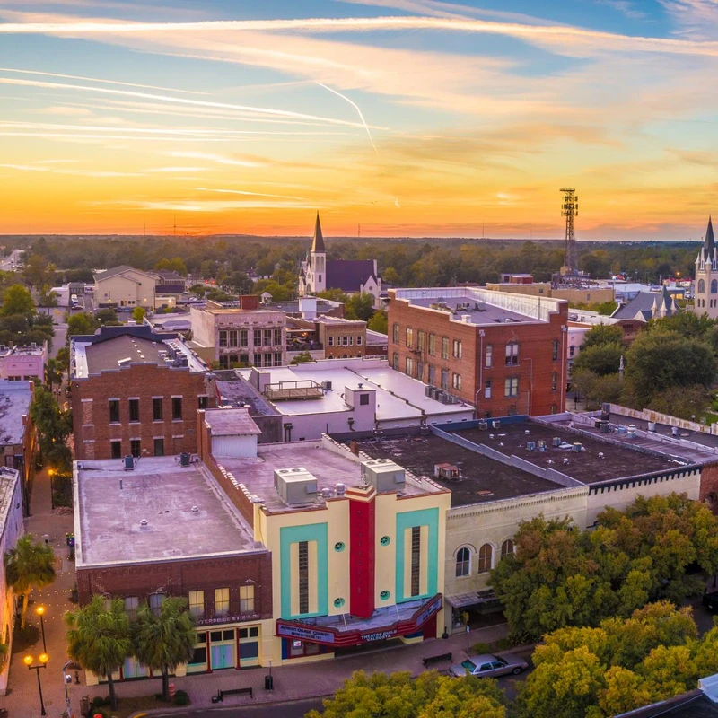 Aerial view of a historic downtown at sunset. A vibrant orange and blue sky sits above classic brick buildings, a courthouse dome, and church steeples.